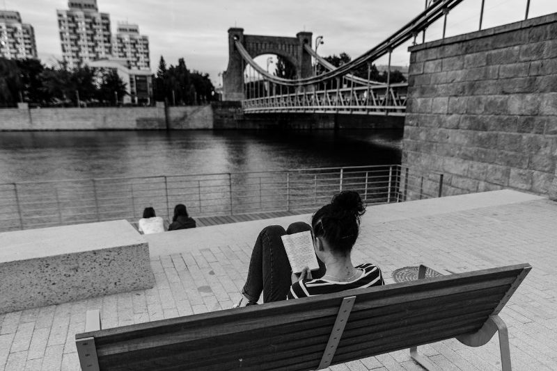 woman reading a book next to a river and bridge in city, black and white photo